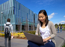 Female Asian student at laptop outside SSA building on Hayward campus