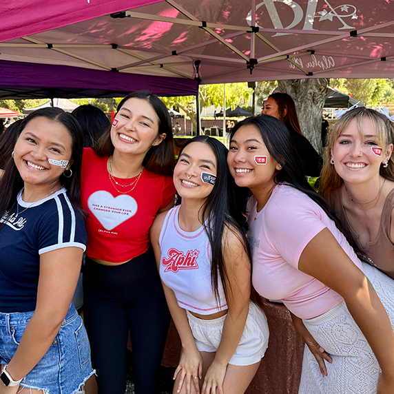 Group of students smiling together at an outdoor campus event, standing under a canopy with stickers on their faces.
