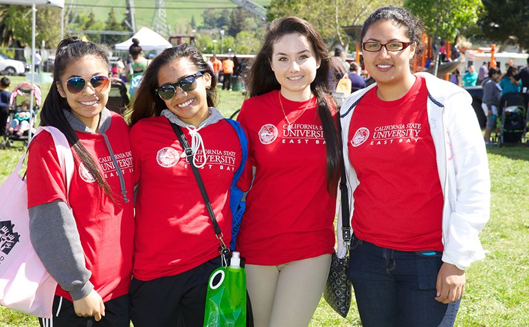 Four female students in red CSUEB shirts gather at an outdoor event