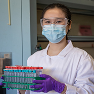 A female in mask and white coat holding test tubes