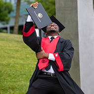 A student in cap in gown on knees holds up degree