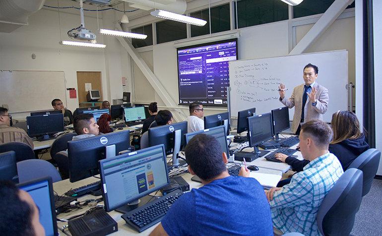 Professor teaches classroom of students with financial ticker in background