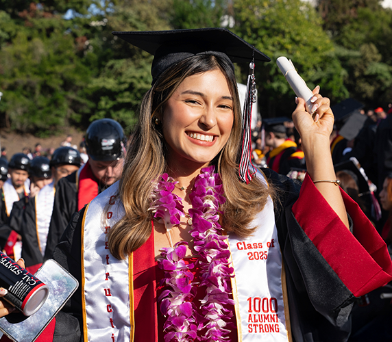 Graduate smiling at commencement while holding a diploma, wearing a cap and gown with a purple lei and a stole that reads ‘Class of 2025,’ with other graduates in the background.