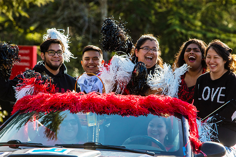 Pioneers cheering as they ride atop a car in a parade