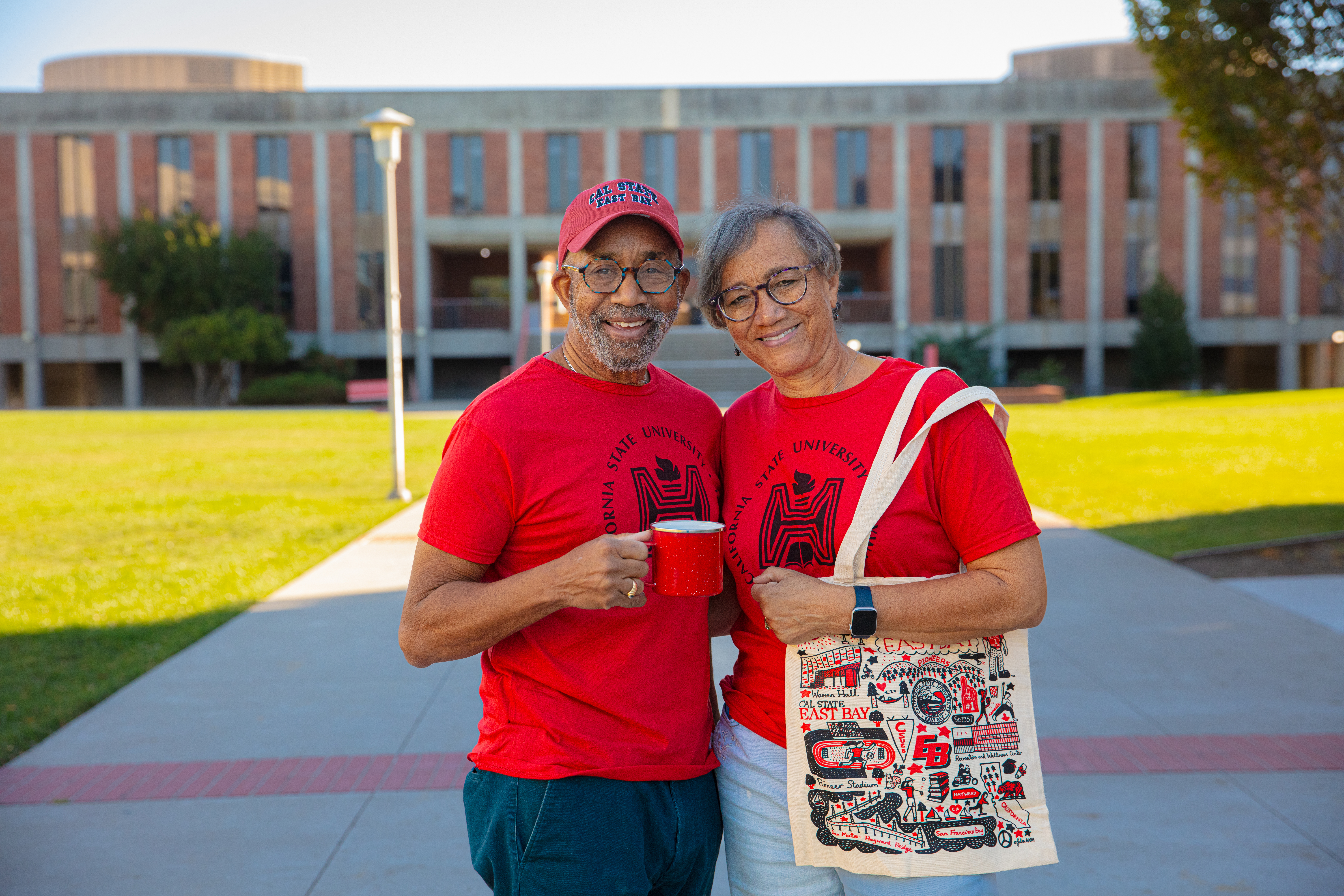 Two alumni wearing Hayward shirts holding coffee mugs in front of a building