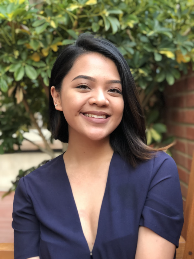 Pilipinx with shoulder length black hair, smiling in front of a brick wall and tree.