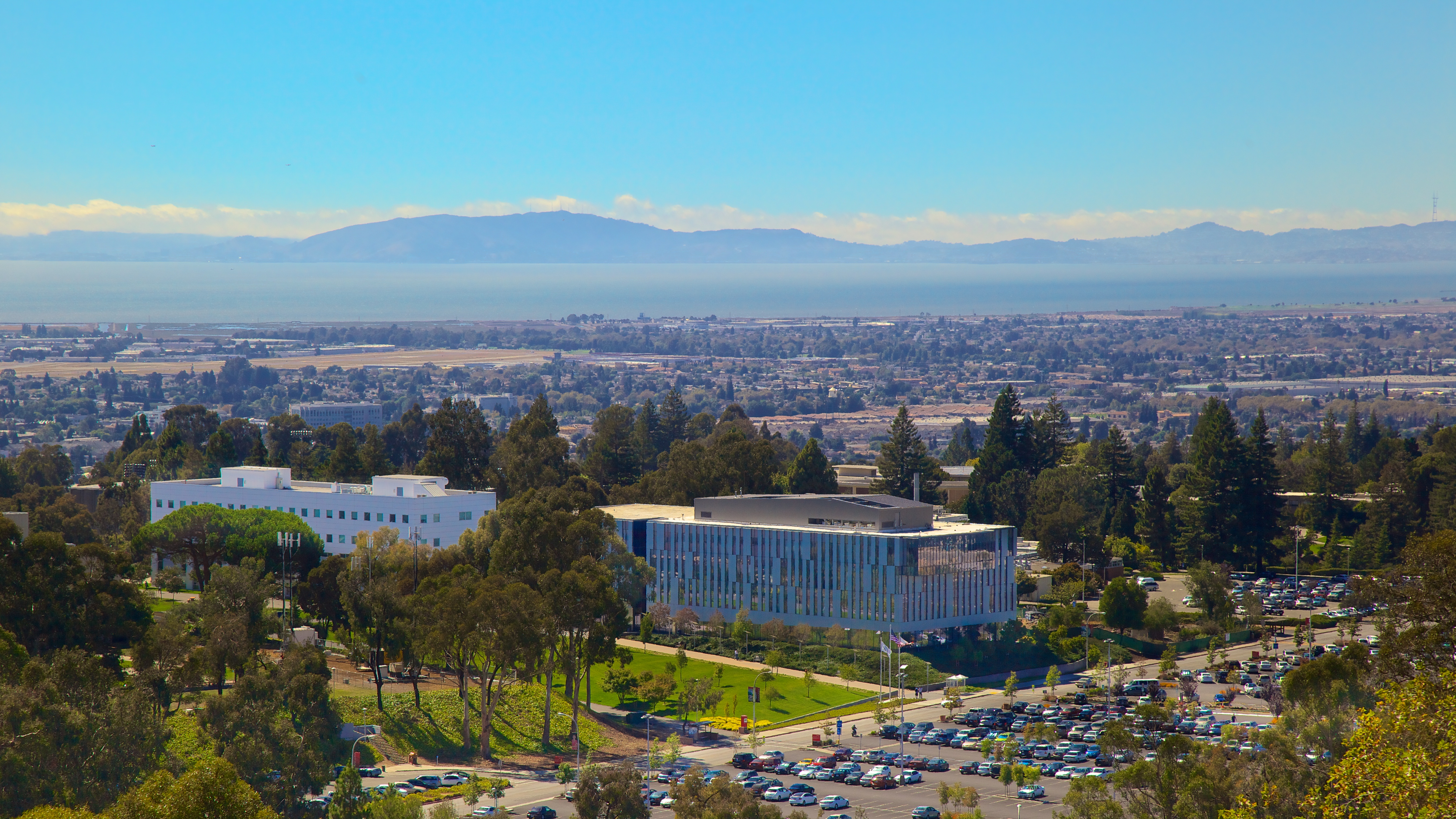 A panoramic view of a campus with modern architecture set against a backdrop of blue waters and mountains in the distance.