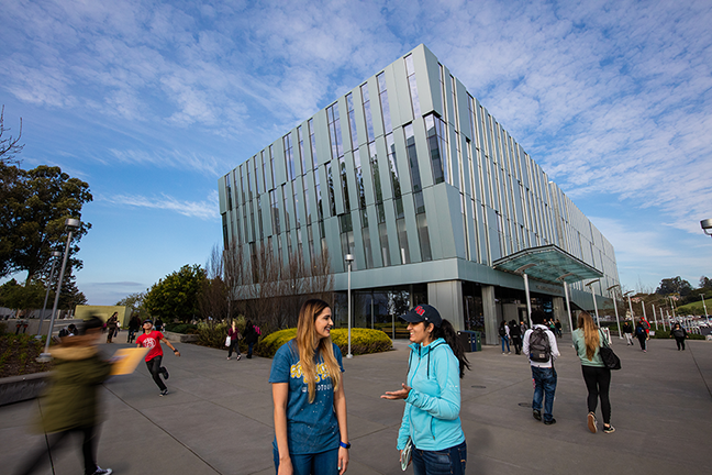 Two students casually converse in front of a modern, glass-fronted academic building on a sunny day.