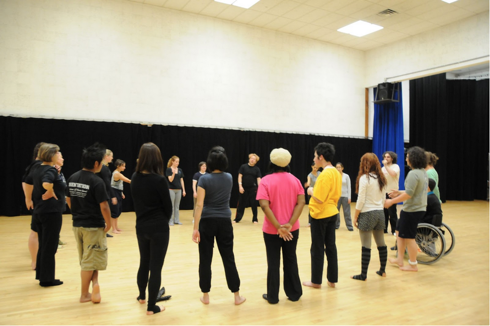 About 25 people of different ages, sizes, shapes, mobilities, and ethnicities gather in a circle in a dance studio, all focused on a conversation together. Photo by: anonymous