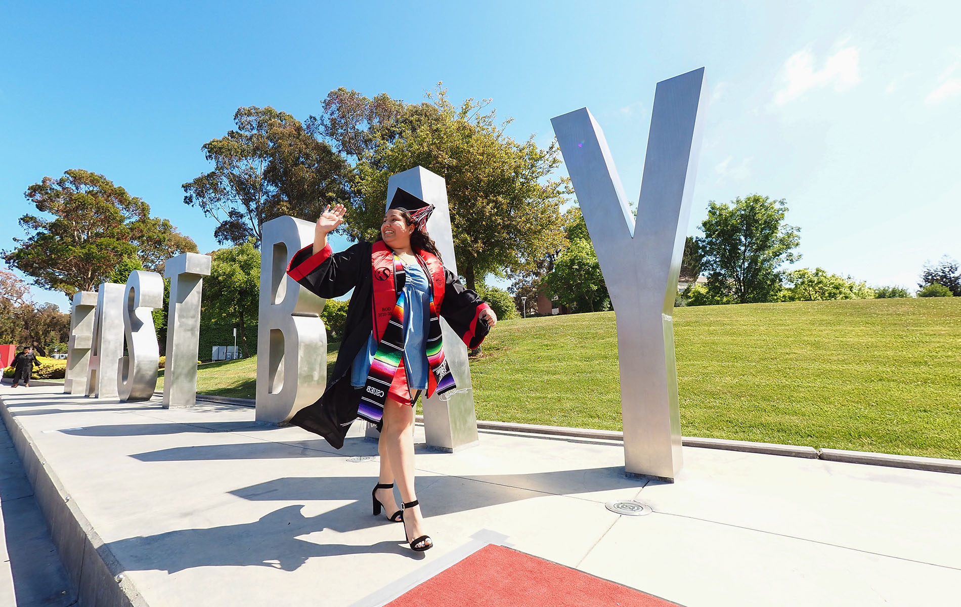 Graduate waves while walking in front of East Bay letters