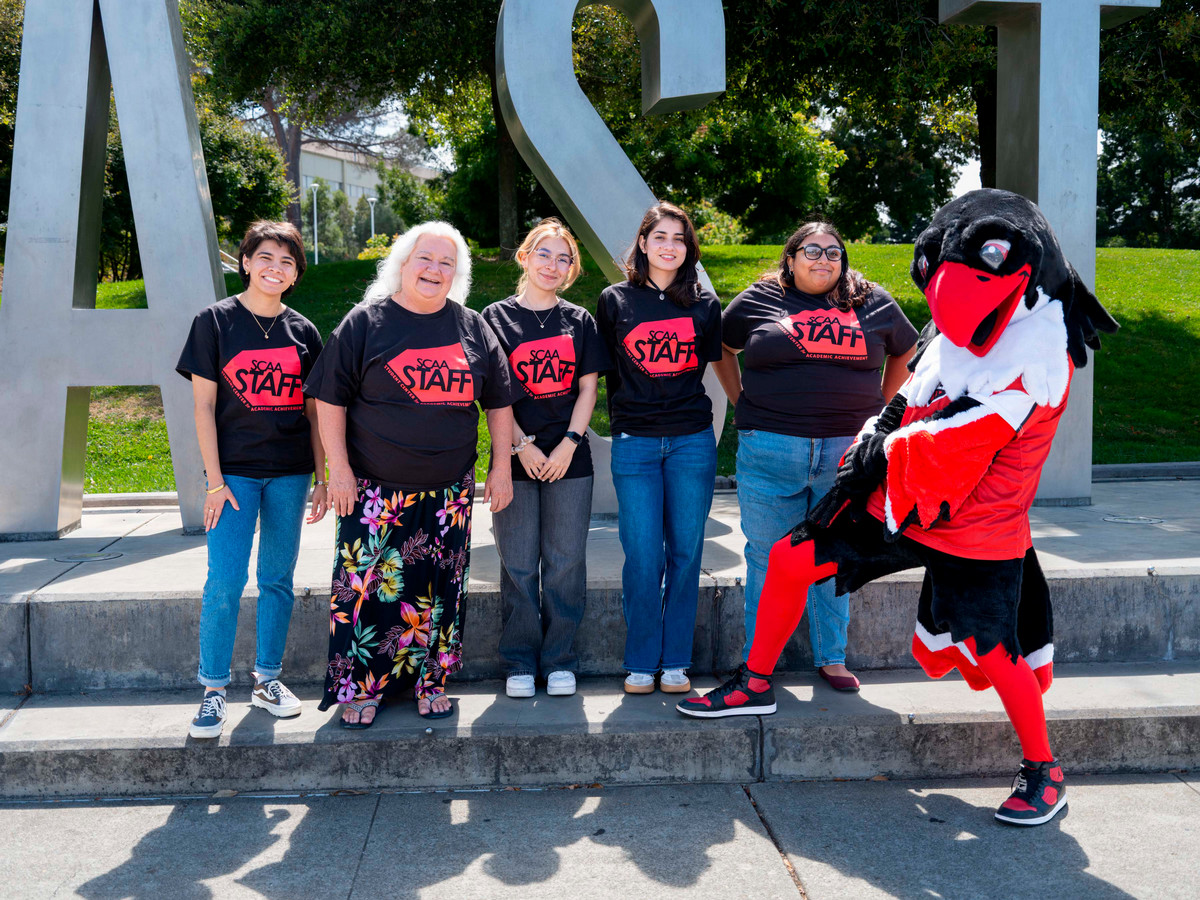 This is a photo of 5 people smiling, posing with the Cal State East Bay mascot on a sunny day