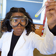A student in a lab coat and goggles holds up a test tube