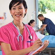 A nurse smiles as another nurse looks at a patient in the background