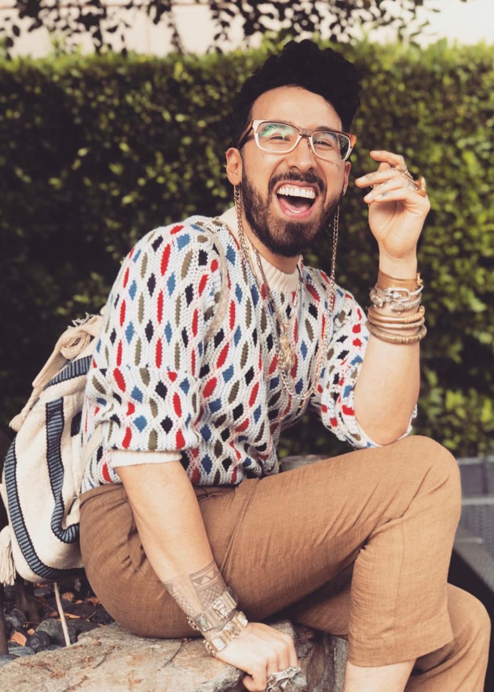 Curly Velasquez smiling wearing a knit sweater, linen pants, and glasses. They are sitting on a stone retaining  wall with a hedge in the background