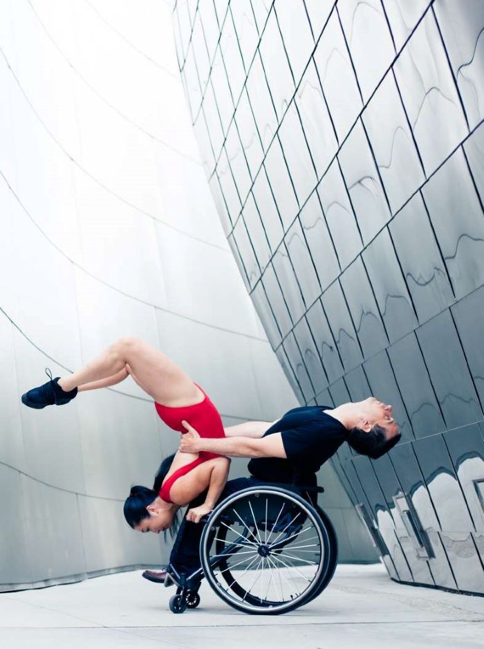 Two dancers in front of the Walt Disney music hall, one dancer using a wheel chair supports his partner as she does a handstand off of his chair armrests.