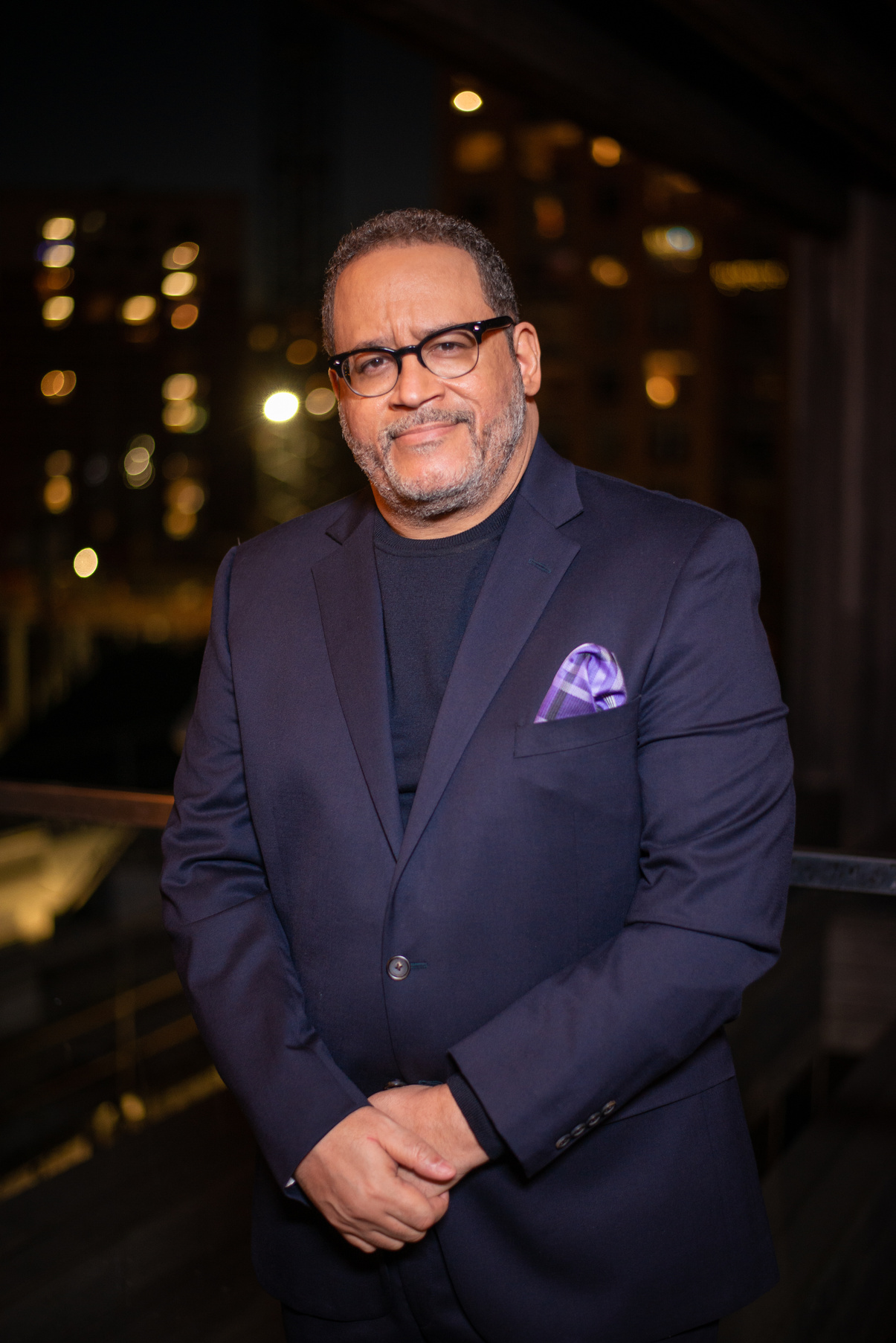 Michael Eric Dyson wearing a navy blazer and horned rimmed glasses looking directly at the camera with his hands clasped in front him.