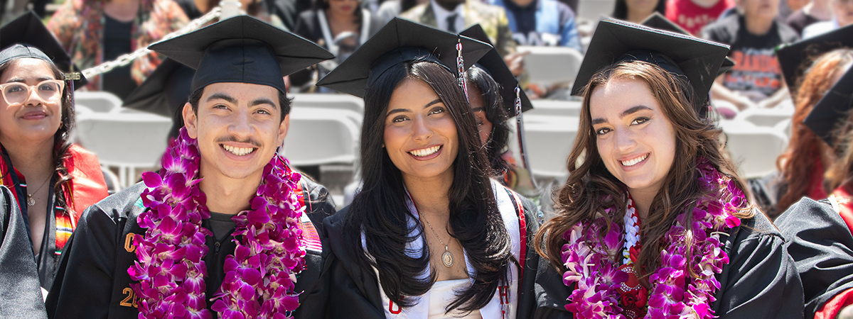 Three graduating students wearing caps, gowns, and purple flower leis smile while seated at an outdoor commencement ceremony.
