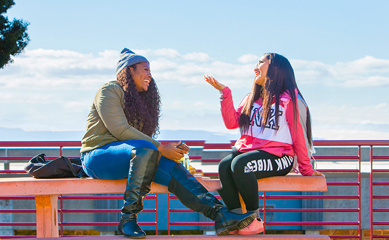 Two students sit together on top of an outdoor picnic table, smiling and talking on a sunny day at Cal State East Bay.