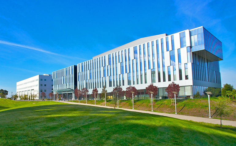 Exterior view of the Student Services and Administration building, surrounded by landscaped lawns, young trees, and a bright blue sky.