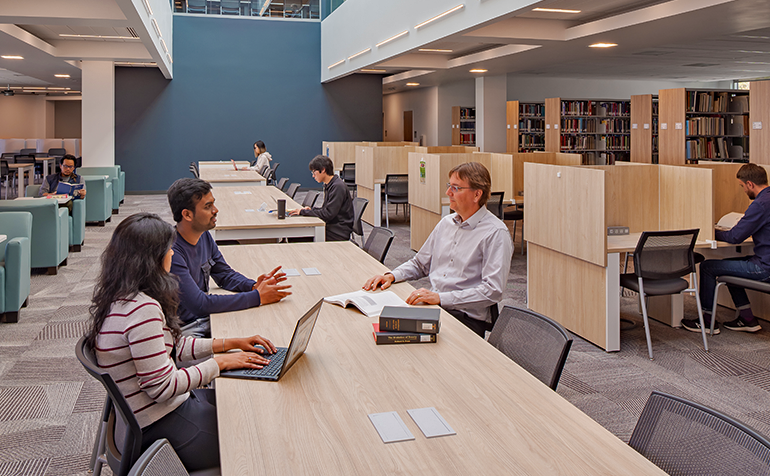 Students and a faculty member study and work at long tables and individual cubicles inside the CORE library, surrounded by bookshelves and soft seating areas.