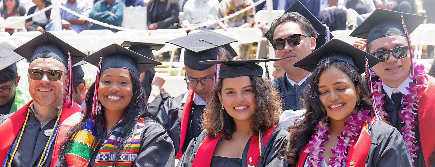 A diverse group of students smiling during their commencement ceremony. They are dressed in their regalia and are wearing various stoles and flower leis.