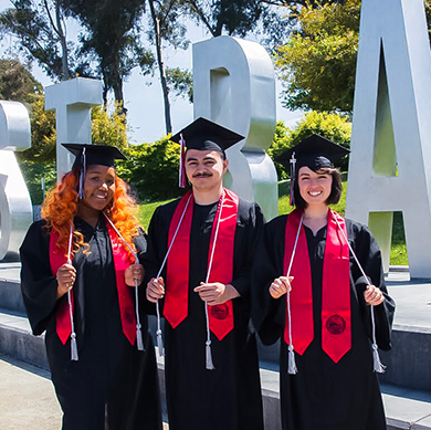  Three students stand in cap and gown in front of the East Bay letters