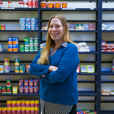  A woman in a blue sweater with long hair stands in front of a shelf stocked with canned goods