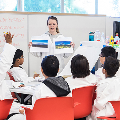  A student teacher in a white coat presents in front of a table of small children wearing white coats