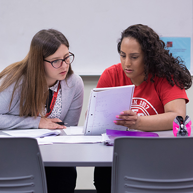  Two female students sitting at desks look over a piece of paper together