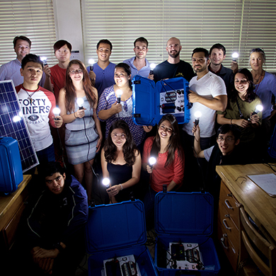  Students and faculty pose in a science lab in the shadows. The only light comes from a blue suitcase being held by a student.