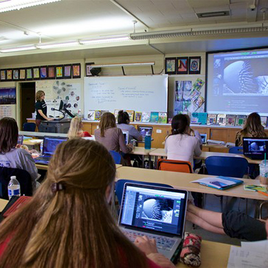  A student works on a laptop while a teacher instructs at the front of a classroom