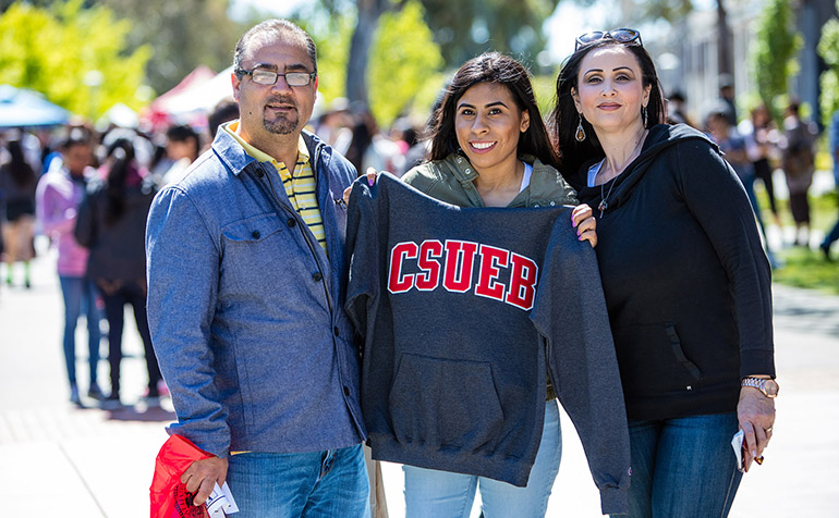 Female student stands with her parents holding a CSUEB sweatshirt