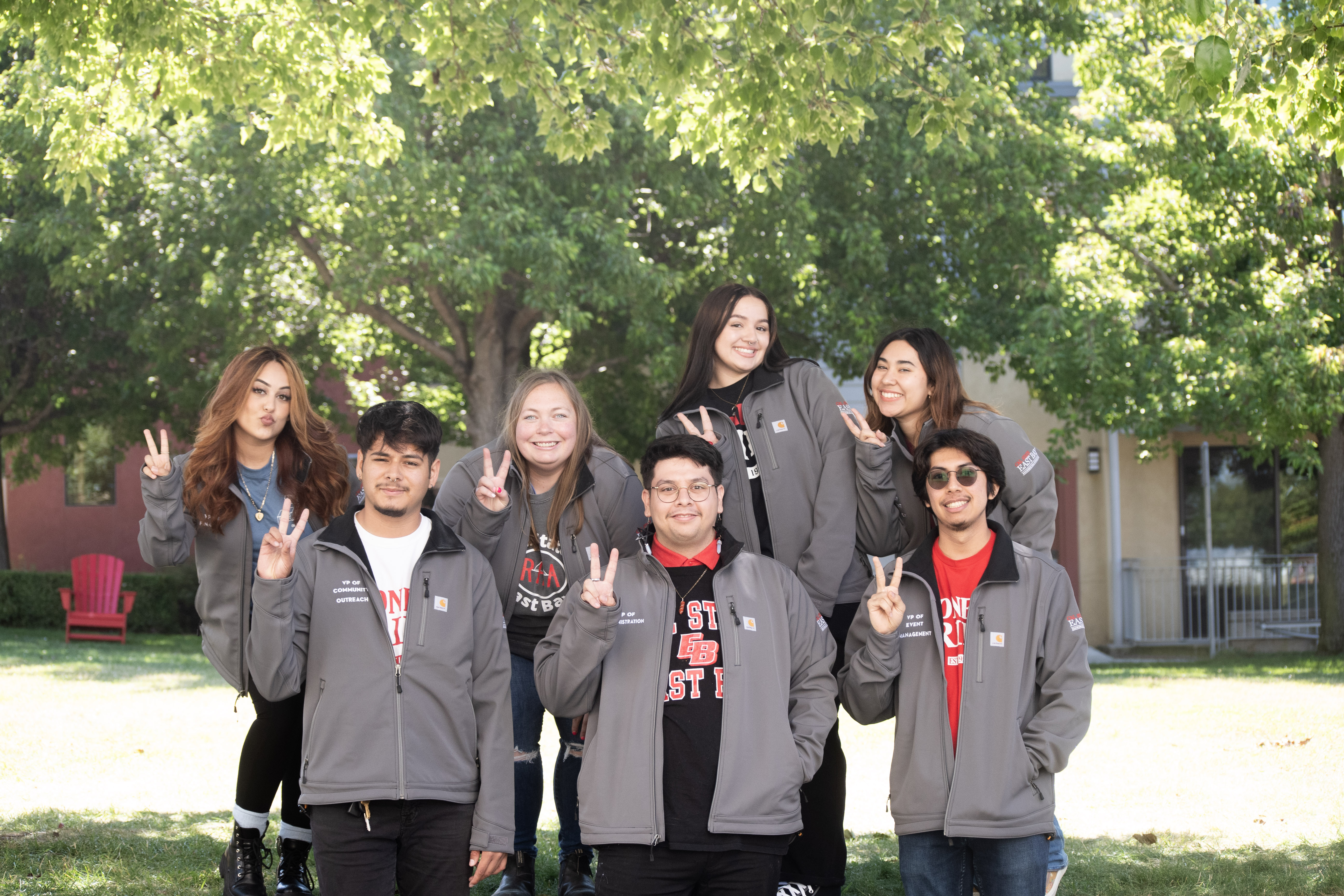 Picture of 5 student leaders wearing Cal State East Bay apparel standing in front of a residence hall. All students are laughing and smiling as four of them lift the fifth one up horizontally in front of them.