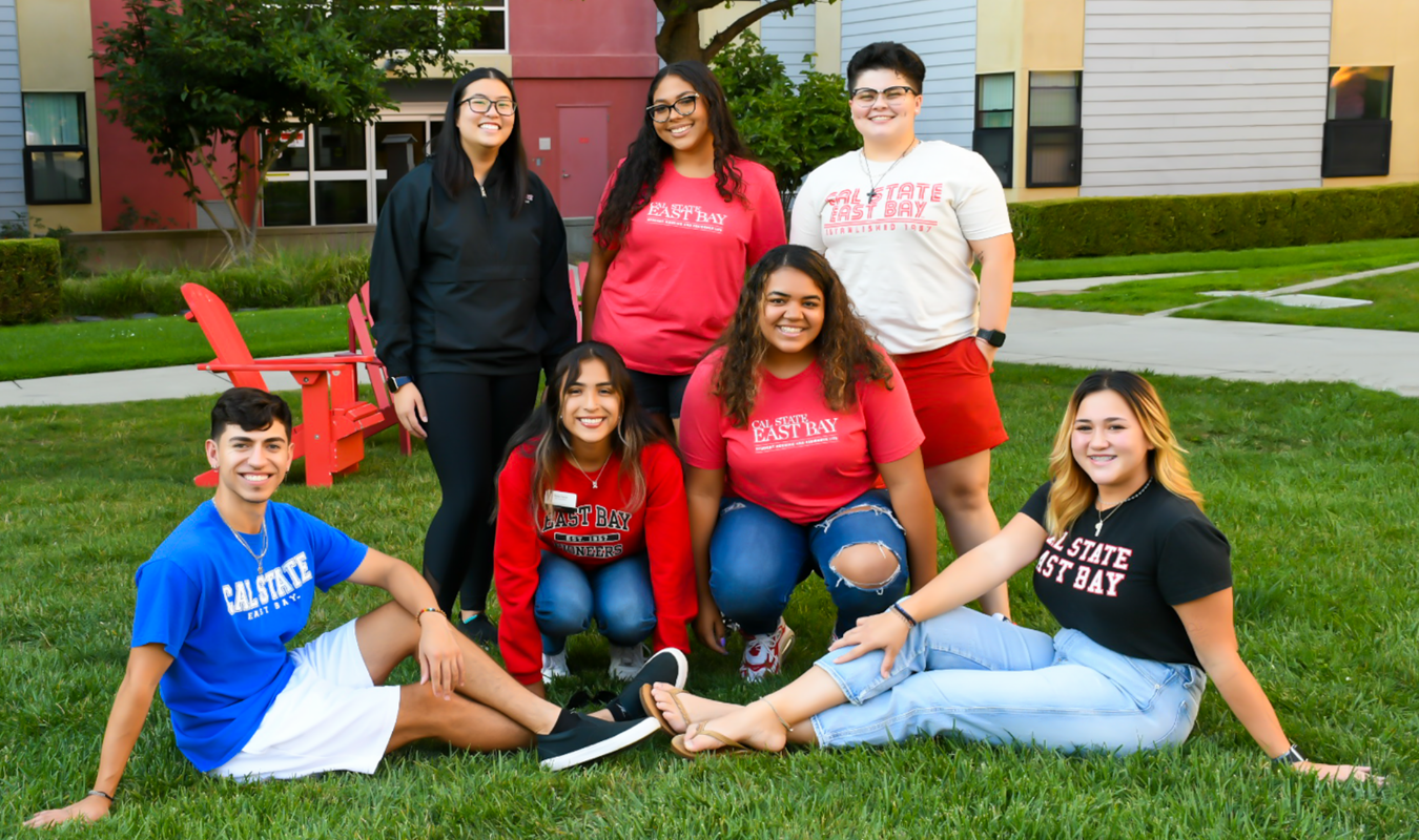 Picture of 7 student leaders wearing Cal State East Bay apparel in front of a residence hall. Two are in front sitting on the grass, two are swatting behind them and the last 3 are standing in the back