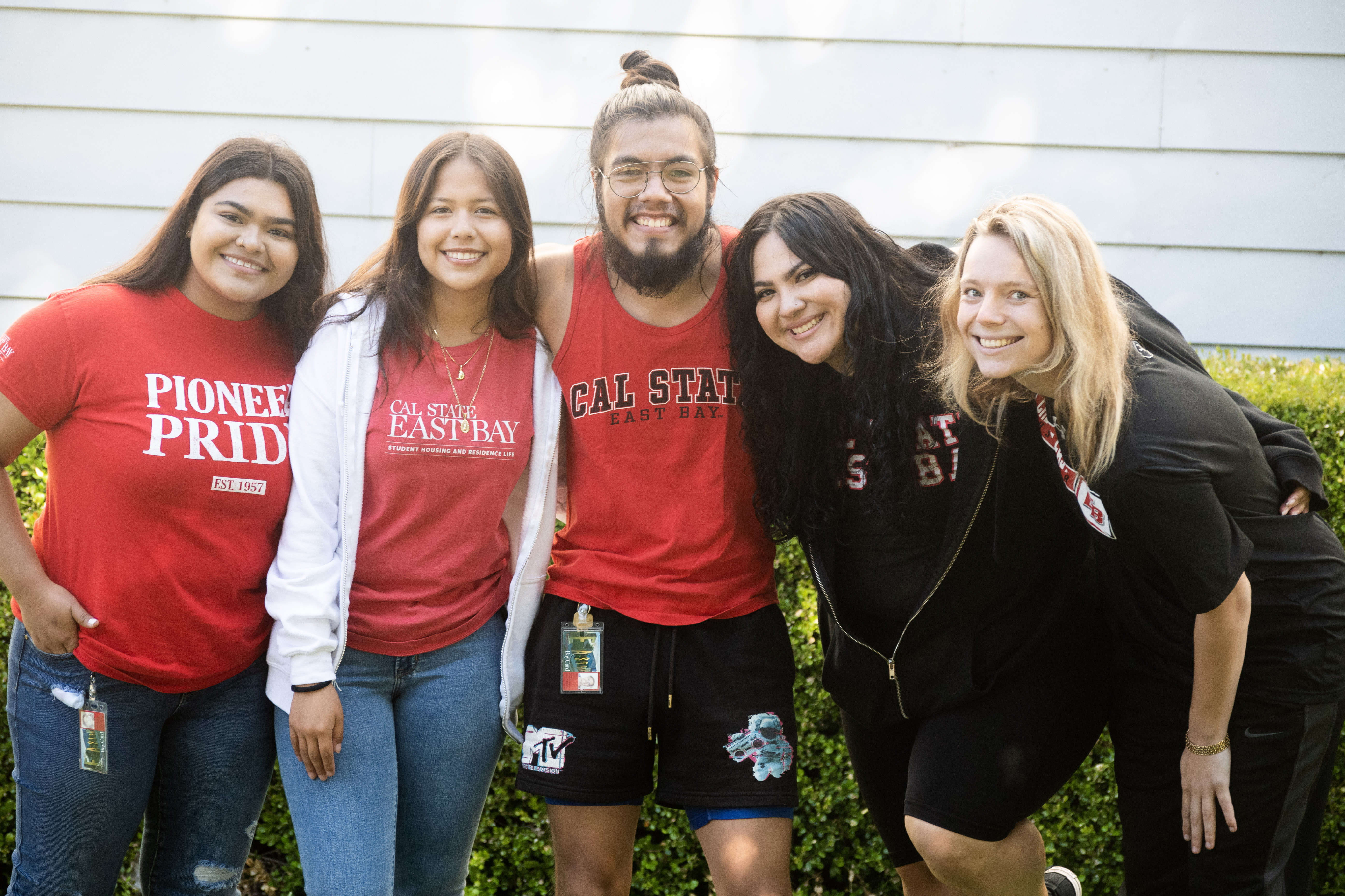 Picture of 4 student leaders wearing Cal State East Bay apparel standing in front of a residence hall with their arms around each other and laughing. 