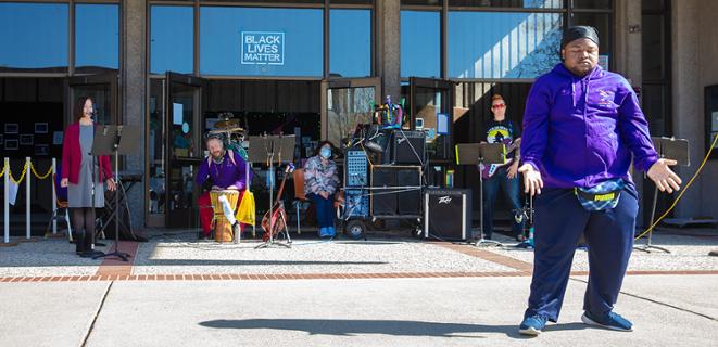 Man wearing purple performs at Coming Alive ceremony while others in background watch 