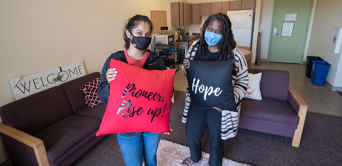 Two women stand in apartment holding pillows