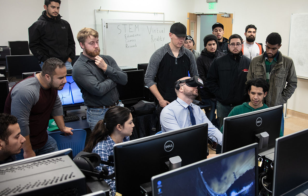 Professor sits at computer wearing VR goggles while several students look on