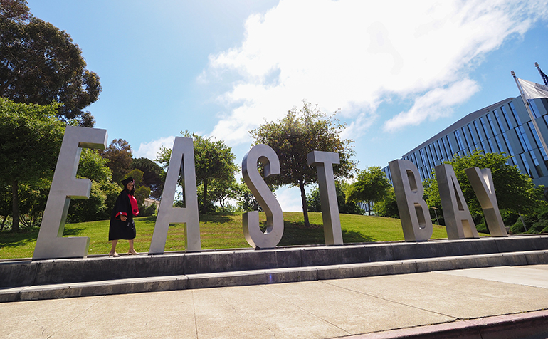 Student graduation gown walks in front of East Bay letters